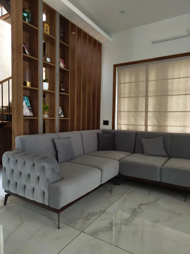 Grey tufted L-shaped sofa beside an oak open-shelf partition and Roman blind